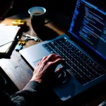 Handen van cybersecurity professional typen op laptop toetsenbord met code op scherm, slot en sleutel op bureau