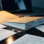 Open auditmap met compliance-checklists, USB-beveiligingssleutel en laptop op donker bureau, verlicht door amberkleurige bureaulamp.