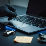 Gehandschoende hand die met een metalen pick een laptoptoetsenbord onderzoekt, blauwe schermgloed op donker bureau met sticky notes en slot.