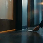 Onbekende hand die een gevouwen briefje onder een gesloten kantoordeur schuift, zwak verlichte gang op de achtergrond.