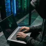 Gehooide ethische hacker typt op laptop in donkere serverruimte, groen sloticoon weerspiegeld op scherm, blauwe serverrackverlichting op de achtergrond.