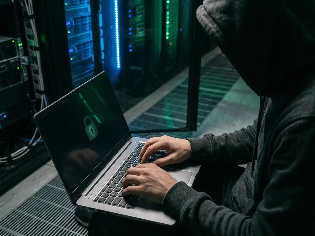 Gehooide ethische hacker typt op laptop in donkere serverruimte, groen sloticoon weerspiegeld op scherm, blauwe serverrackverlichting op de achtergrond.