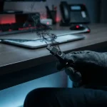 Gehandschoende hand houdt een gebarsten USB-stick vast waaruit donkere rook kringelt, met een laptop op de achtergrond.
