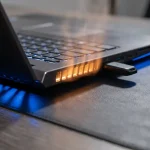 Oververhitte laptop op donker bureau met zichtbare warmtegloed en verborgen USB-apparaat onder toetsenbordrand.