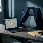 Laptop met verdachte e-mail op bureau naast bedrijfspas en envelop, met een pak over de stoel in een modern kantoor.