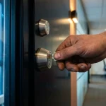 Verweerde hand die een sleutel in een modern deurslot steekt, met een stalen grendel op de achtergrond in een kantoorgang.