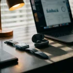 Smartwatch en draadloze oordopjes op een modern bureau naast een open laptop, met dramatische schaduwen en staalblauwe tinten.