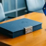 Gesloten overheids­compliance­map met metalen sluiting op modern bureau naast een EU-vlag, koele blauwtinten en warm amberkleurig licht.